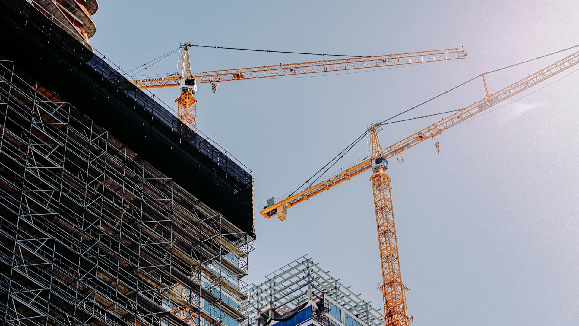 Twee gele torenkranen boven een hoogbouwconstructie met steigers en staalstructuur onder een heldere lucht.